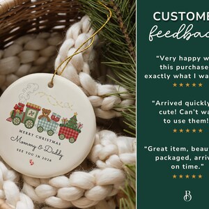 May include: A round Christmas ornament with a train design. The train cars are decorated with a teddy bear, gifts, and a Christmas tree. The text reads "Merry Christmas Mommy & Daddy See You in 2026". The ornament is cream-colored with a gold string.