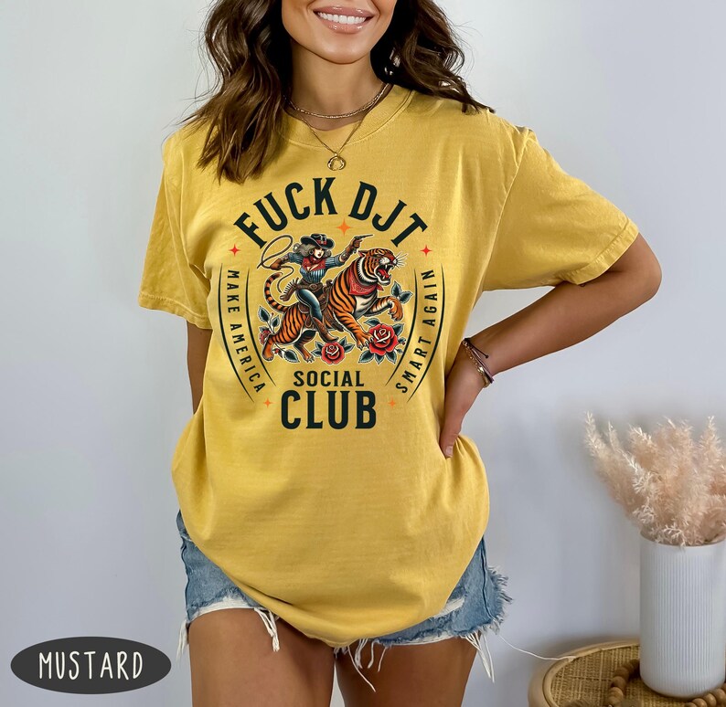 May include: A mustard yellow t-shirt with a graphic print of a woman riding a tiger. The graphic includes the text "F*CK DJT" in a circle with the words "Make America Smart Again" and "Social Club".