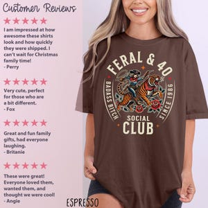 May include: Espresso-colored t-shirt featuring a graphic of a woman riding a tiger, with the text "FERAL & 40" and "BADASS BITCH SOCIAL CLUB SINCE 1986". The shirt has a crew neck and short sleeves.