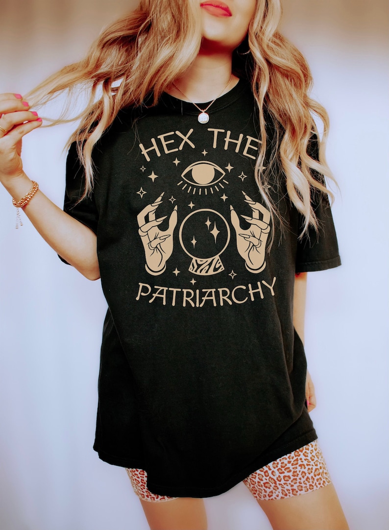 May include: Black t-shirt with a gold graphic of a hand holding a crystal ball with an eye above it. The text "Hex the Patriarchy" is printed below the graphic.