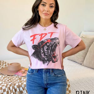 Peut inclure: T-shirt rose avec un motif noir et blanc d'un tigre rugissant avec des accents rouges. Le motif comprend le texte "FDT".