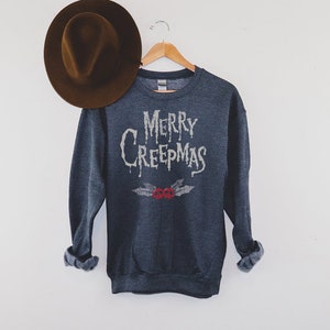 May include: Dark gray sweatshirt with the text "MERRY CREEPMAS" in a dripping font, with a pair of red skull holly berries. A brown fedora hat rests on the left side. The sweatshirt is hanging on a wooden hanger.