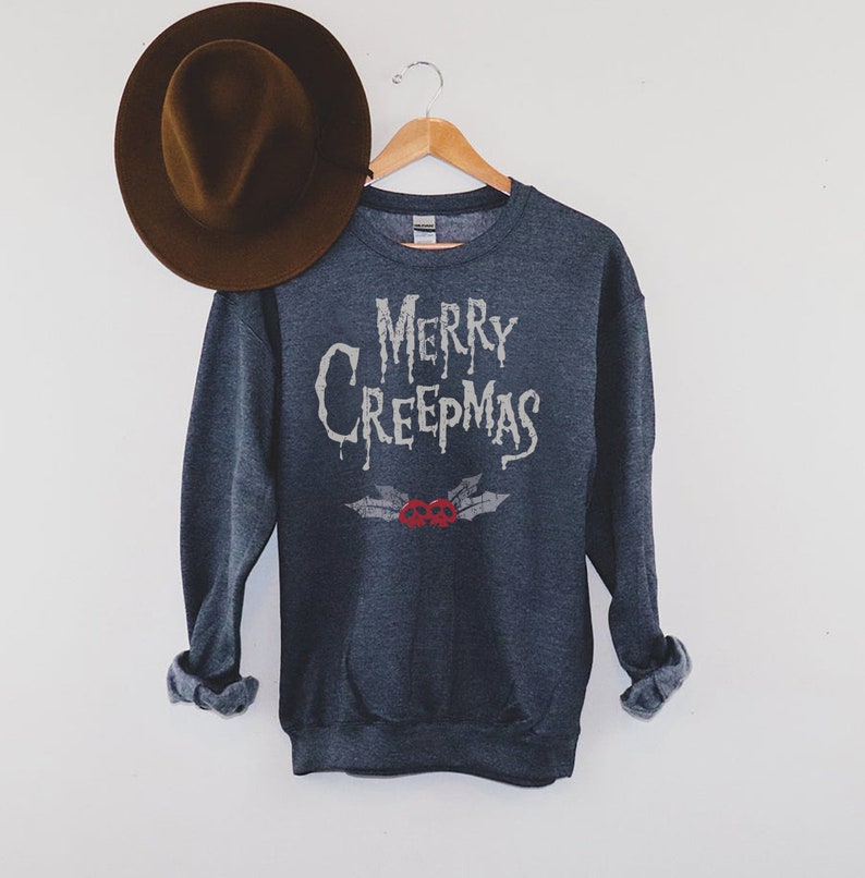 May include: Dark gray sweatshirt with the text "MERRY CREEPMAS" in a dripping font, with a pair of red skull holly berries. A brown fedora hat rests on the left side. The sweatshirt is hanging on a wooden hanger.