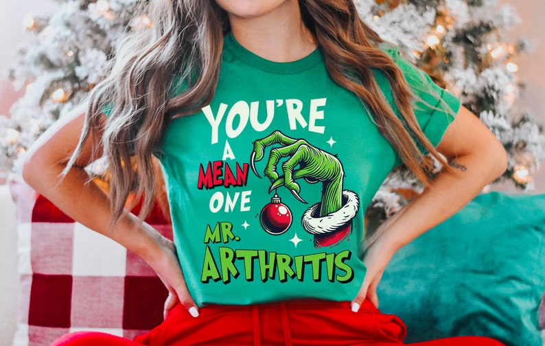 K&ouml;nnte beinhalten: Gr&uuml;nes T-Shirt mit einer Grinch-&auml;hnlichen Hand, die einen roten Weihnachtsbaumschmuck h&auml;lt. Der Text auf dem Shirt lautet "You're a mean one Mr. Arthritis".