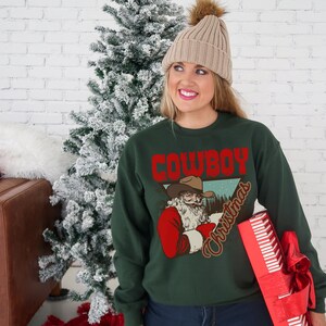 May include: A green Christmas sweater with a vintage illustration of Santa Claus wearing a cowboy hat and the text "Cowboy Christmas".