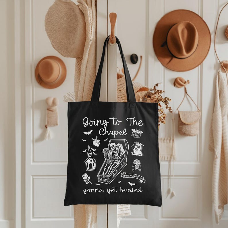 May include: Black tote bag with the text "Going to The Chapel gonna get buried" and illustrations of skeletons, bats, and a church. The bag is hanging on a wooden hook.