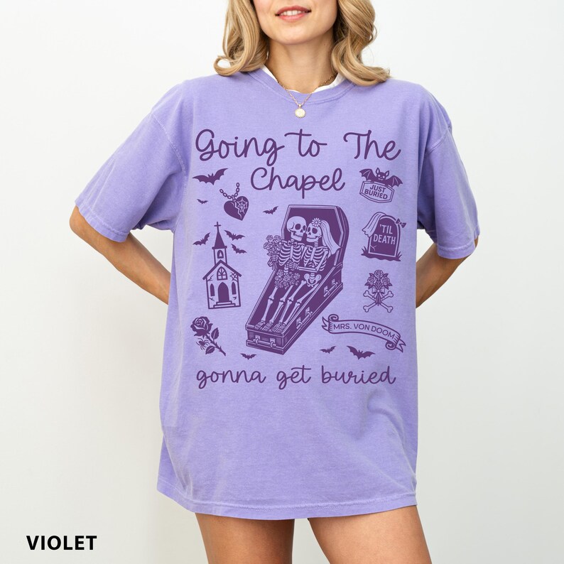 May include: Violet-colored t-shirt with the text "Going to The Chapel" and a graphic of skeletons in a coffin. Additional graphics include bats, a church, and a rose. The text "gonna get buried" is also included.