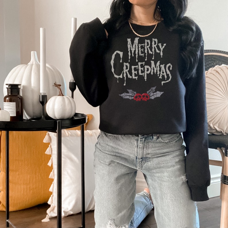 May include: Black sweatshirt with the text "MERRY CREEPMAS" in a spooky font, with a design of skulls and holly leaves. The sweatshirt is a crew neck style. The image also shows white pumpkins and candles.