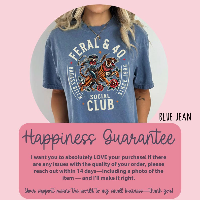 May include: Blue jean colored t-shirt with a graphic featuring a woman riding a tiger, roses, and the text "FERAL & 40 BADASS BITCH SOCIAL CLUB SINCE 1986". The shirt has a crew neck and short sleeves.