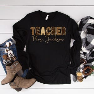 May include: Black long-sleeve shirt with the word "TEACHER" in a brown leopard print design and the name "Mrs. Jackson" in a gold script. The shirt is paired with denim jeans, brown boots, and a black and white scarf.