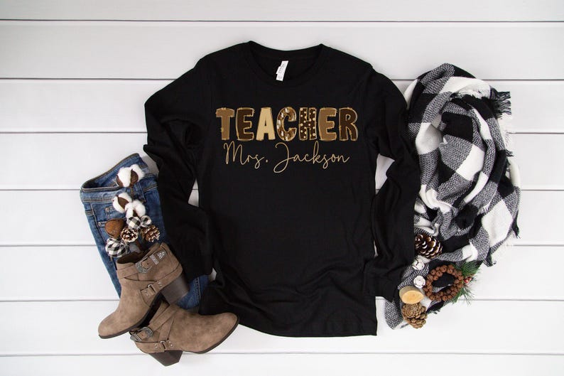 May include: Black long-sleeve shirt with the word "TEACHER" in a brown leopard print design and the name "Mrs. Jackson" in a gold script. The shirt is paired with denim jeans, brown boots, and a black and white scarf.