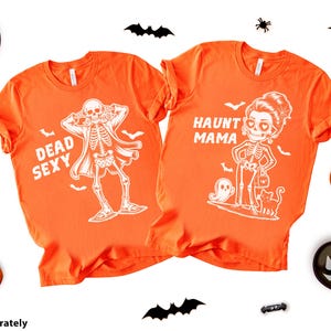 K&ouml;nnte beinhalten: Zwei orangefarbene T-Shirts mit Halloween-Designs. Eines zeigt ein Skelett und den Text "Dead Sexy". Das andere sagt "Haunt Mama" mit einer Skelettfrau, einem Geist und einer Katze. Halloween-Dekorationen umgeben die Shirts.