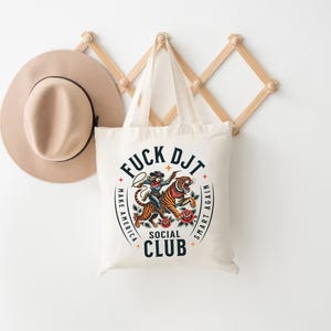 May include: A white canvas tote bag with a graphic design featuring a tiger and a person riding it. The text on the bag reads "F*CK DJT MAKE AMERICA SMART AGAIN SOCIAL CLUB".