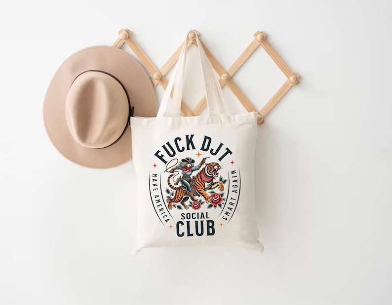 May include: A white canvas tote bag with a graphic design featuring a tiger and a person riding it. The text on the bag reads "F*CK DJT MAKE AMERICA SMART AGAIN SOCIAL CLUB".