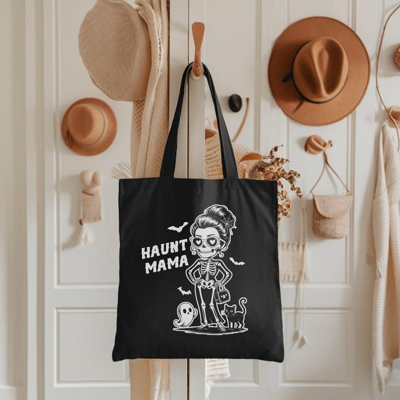 K&ouml;nnte beinhalten: Schwarze Tragetasche mit dem Schriftzug "HAUNT MAMA" und einer wei&szlig;en Grafik einer Skelettfrau, eines Geistes, einer schwarzen Katze und Flederm&auml;usen. Die Tragetasche hat schwarze Griffe und h&auml;ngt an einem Holzhaken.