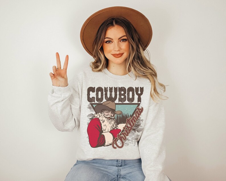 May include: A gray sweatshirt with a vintage-style graphic of a cowboy Santa Claus. The graphic features the text "Cowboy Christmas" in a brown, white, and blue color scheme.