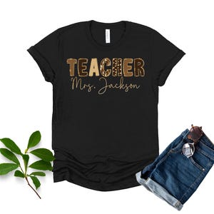 May include: Black t-shirt with the word "TEACHER" in a brown leopard print design and the name "Mrs. Jackson" in gold script. The shirt is paired with denim shorts and sunglasses.