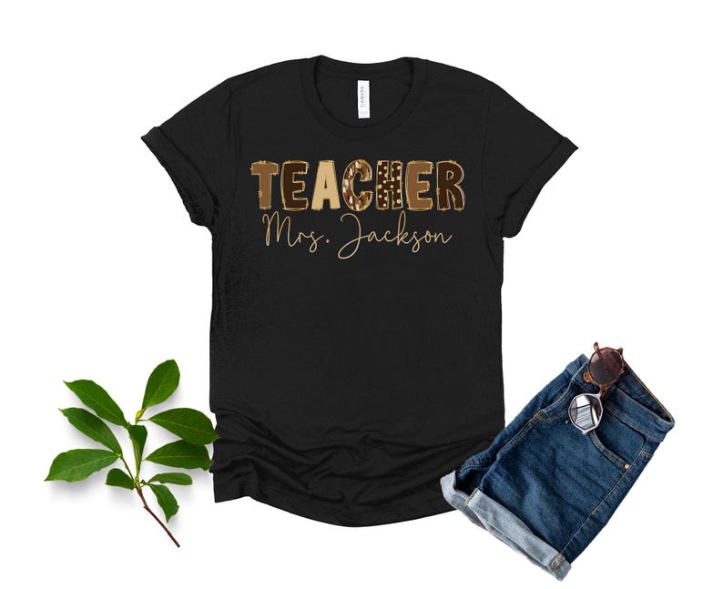 May include: Black t-shirt with the word "TEACHER" in a brown leopard print design and the name "Mrs. Jackson" in gold script. The shirt is paired with denim shorts and sunglasses.