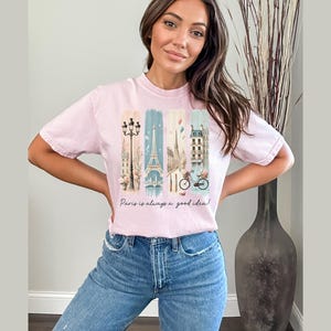 May include: A pink t-shirt with a graphic print of Parisian landmarks, including the Eiffel Tower, a street lamp, and a bicycle. The text "Paris is always a good idea!" is printed below the graphic.