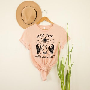 May include: A peach-colored t-shirt with a black graphic design featuring a crystal ball, two hands, and an eye. The text on the shirt reads "Hex the Patriarchy".