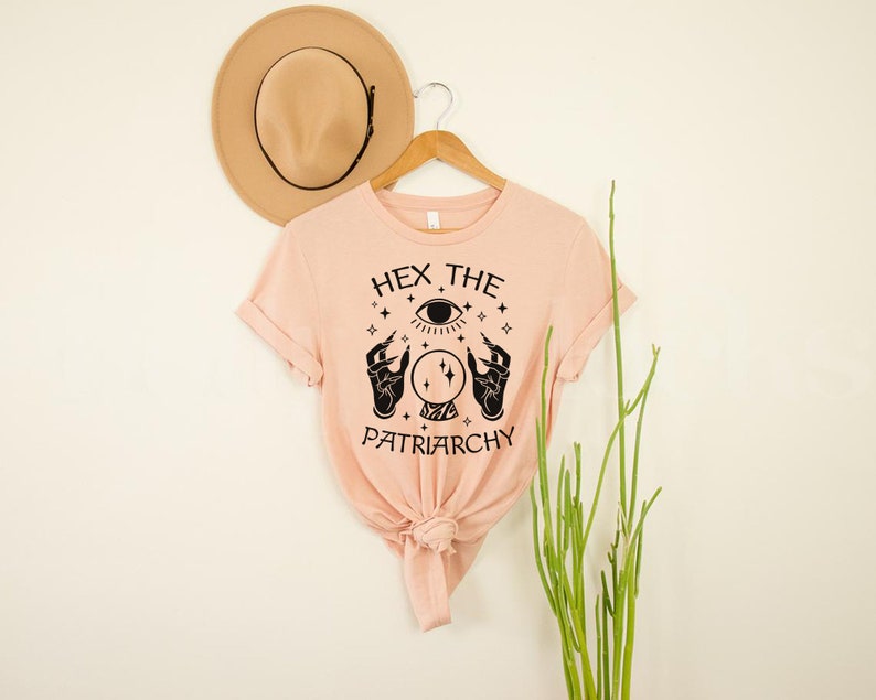 May include: A peach-colored t-shirt with a black graphic design featuring a crystal ball, two hands, and an eye. The text on the shirt reads "Hex the Patriarchy".