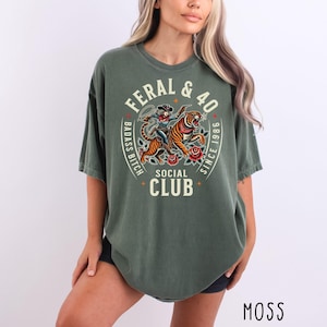 May include: Moss green t-shirt with a graphic design featuring a tiger, a woman with a lasso, and the text "FERAL & 40 BADASS BITCH SOCIAL CLUB SINCE 1986". The shirt has a relaxed fit.