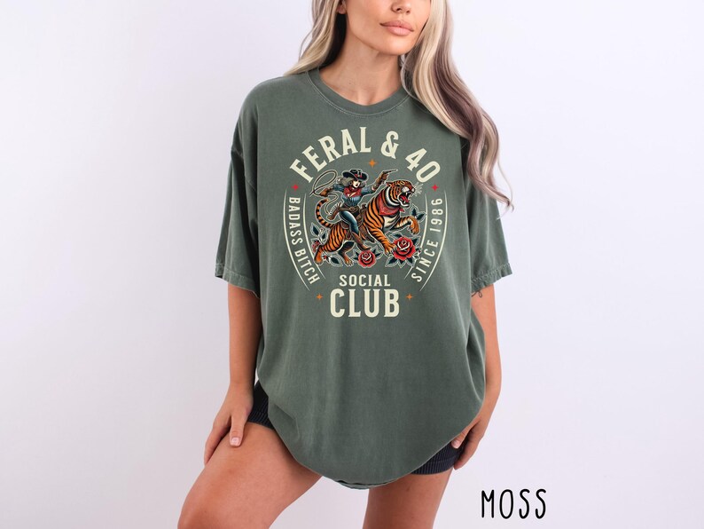 May include: Moss green t-shirt with a graphic design featuring a tiger, a woman with a lasso, and the text "FERAL & 40 BADASS BITCH SOCIAL CLUB SINCE 1986". The shirt has a relaxed fit.