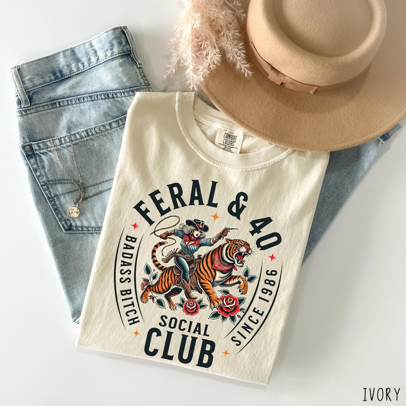 May include: Ivory-colored t-shirt with a graphic of a cowgirl riding a tiger, surrounded by text that reads "FERAL & 40 BADASS BITCH SOCIAL CLUB SINCE 1986." Light blue jeans, a tan hat, and a necklace are also in the image.