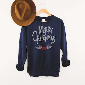 May include: Navy blue sweatshirt with the text "MERRY CREEPMAS" in a dripping font. Below the text is a design of holly leaves and two red skulls. A brown hat hangs in the background.