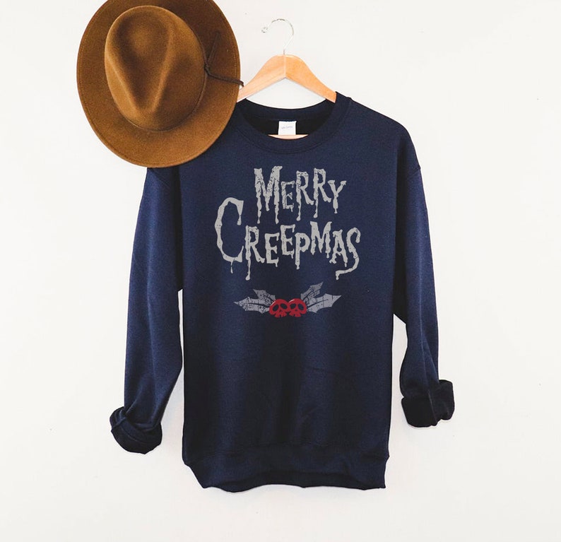 May include: Navy blue sweatshirt with the text "MERRY CREEPMAS" in a dripping font. Below the text is a design of holly leaves and two red skulls. A brown hat hangs in the background.