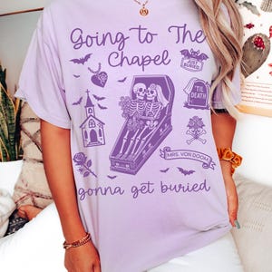 May include: Orchid-colored t-shirt with a gothic wedding theme. The shirt features the text "Going to The Chapel" and "gonna get buried" with illustrations of bats, a chapel, and skeletons in a coffin. The word "ORCHID" is printed at the bottom.