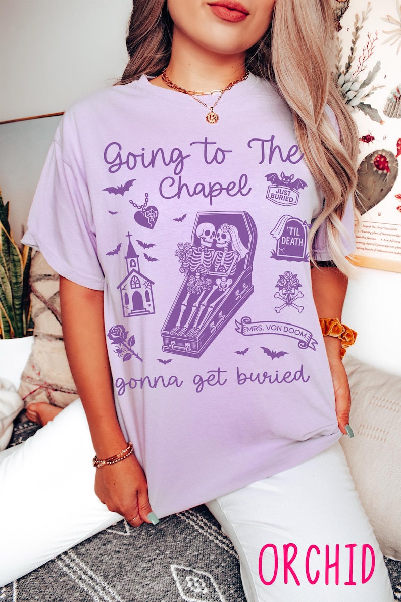 May include: Orchid-colored t-shirt with a gothic wedding theme. The shirt features the text "Going to The Chapel" and "gonna get buried" with illustrations of bats, a chapel, and skeletons in a coffin. The word "ORCHID" is printed at the bottom.