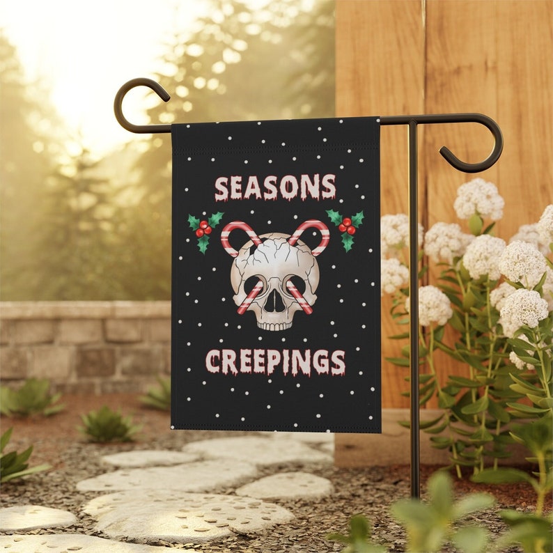 Puede incluir: Una bandera de jard&iacute;n negra con lunares blancos presenta una calavera con ojos de bast&oacute;n de caramelo y acentos de acebo. Las palabras "SEASONS CREEPINGS" est&aacute;n impresas en una fuente espeluznante. La bandera se muestra en un soporte de metal negro.