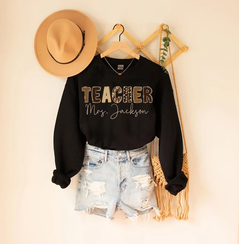 May include: Black sweatshirt with the word "TEACHER" in a patterned design and the name "Mrs. Jackson" in script. The sweatshirt is paired with denim shorts and a tan hat. A woven bag hangs nearby.