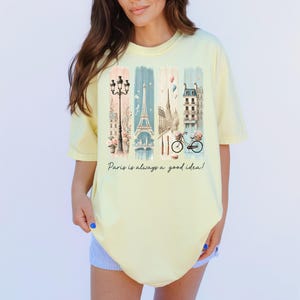 May include: A yellow t-shirt with a graphic print of a Parisian street scene, including the Eiffel Tower, a bicycle, and a street lamp. The text "Paris is always a good idea!" is printed below the graphic.