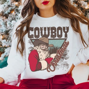May include: White sweatshirt with a vintage-style graphic of a cowboy Santa Claus wearing a hat and holding a red bag. The graphic is in a triangle shape with the words "Cowboy Christmas" written in a distressed font.
