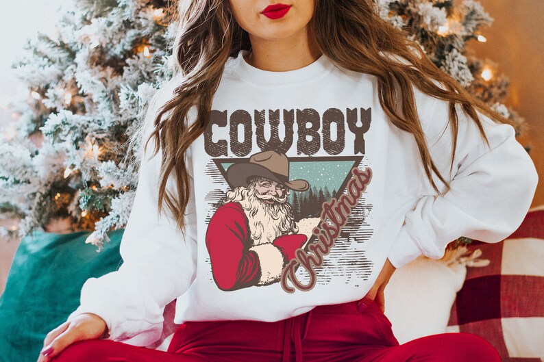 May include: White sweatshirt with a vintage-style graphic of a cowboy Santa Claus wearing a hat and holding a red bag. The graphic is in a triangle shape with the words "Cowboy Christmas" written in a distressed font.