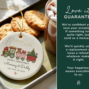May include: A white ceramic Christmas ornament with a train design. The train cars feature a Christmas tree, a teddy bear, and gifts. The text reads "Merry Christmas Mommy & Daddy See You in 2026". A gold string is attached for hanging.