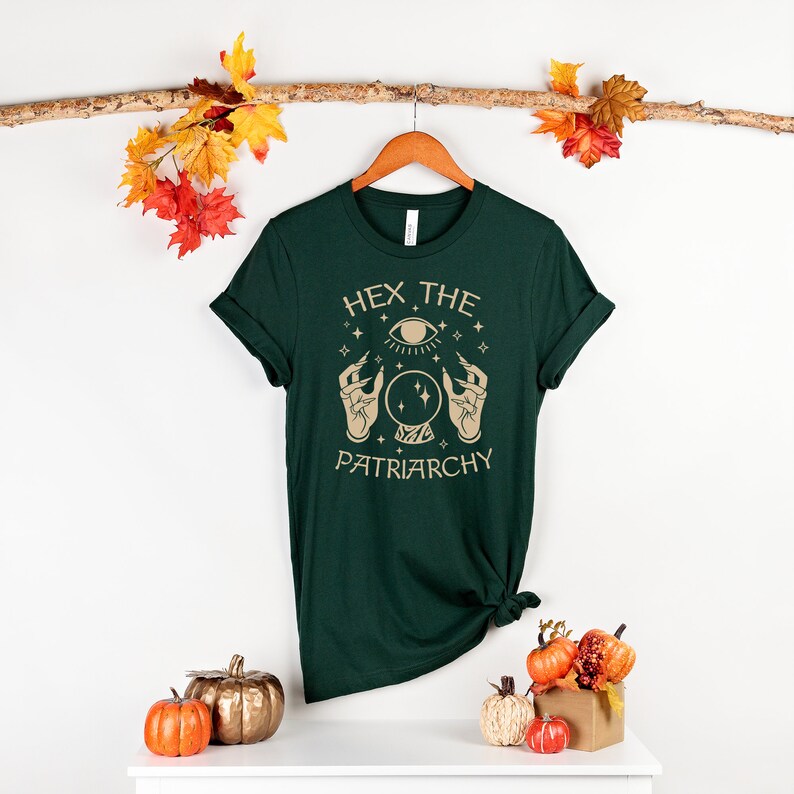 May include: A dark green t-shirt with a gold graphic of a hand holding a crystal ball with an eye inside. The text "Hex the Patriarchy" is printed below the graphic.