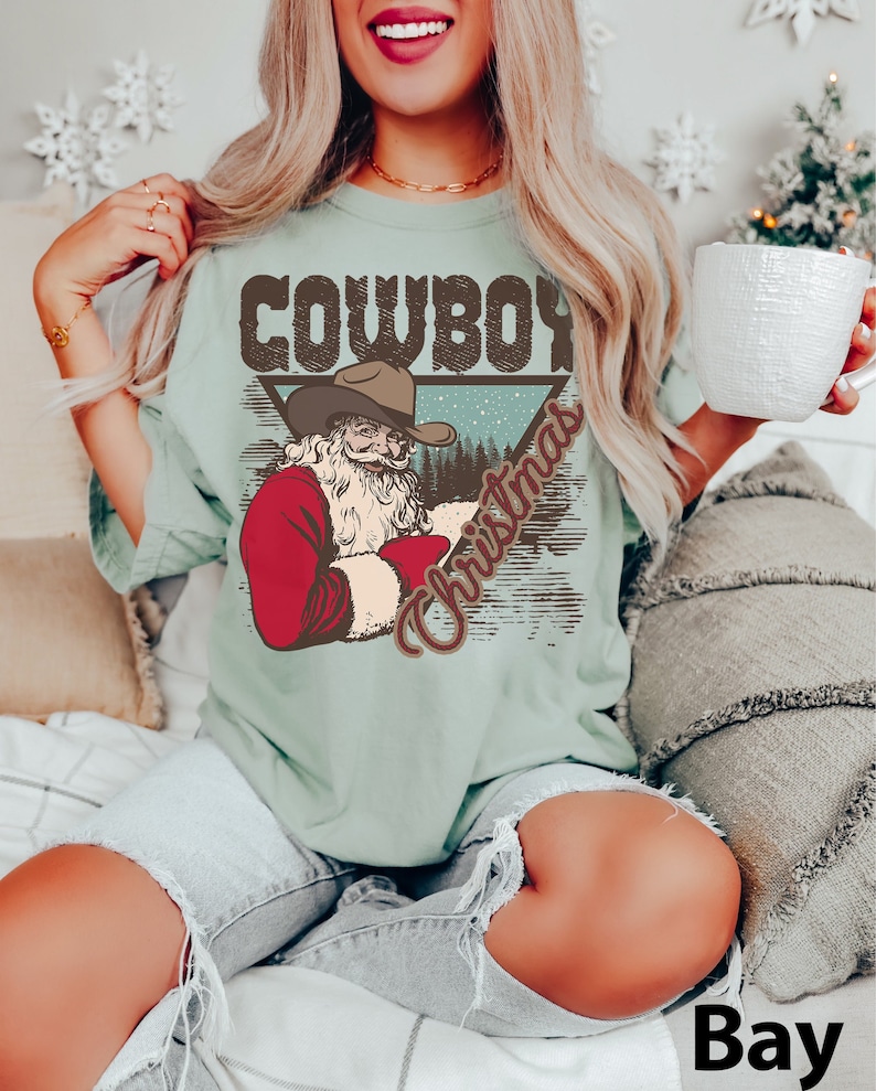 May include: A light blue t-shirt with a vintage-style graphic of Santa Claus wearing a cowboy hat. The graphic says "Cowboy Christmas" in brown text.