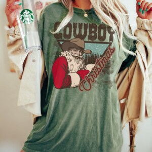 May include: Green t-shirt with a vintage-style graphic of a cowboy Santa Claus with the text "Cowboy Christmas".