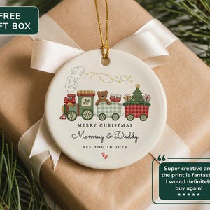 May include: A white ceramic Christmas ornament featuring a train design with a teddy bear, gifts, and a Christmas tree. The ornament has the text "MERRY CHRISTMAS Mommy & Daddy SEE YOU IN 2026". The ornament is on a wrapped gift box.