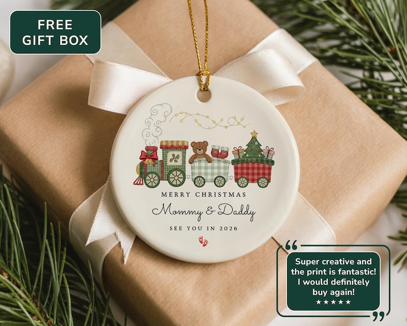May include: A white ceramic Christmas ornament featuring a train design with a teddy bear, gifts, and a Christmas tree. The ornament has the text "MERRY CHRISTMAS Mommy & Daddy SEE YOU IN 2026". The ornament is on a wrapped gift box.