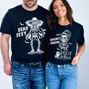 K&ouml;nnte beinhalten: Schwarze T-Shirts mit Halloween-Skelett-Designs. Eines zeigt ein Skelett mit Mantel und dem Text "DEAD SEXY". Das andere zeigt ein Skelett mit Handtasche, Geist und Katze mit dem Text "HAUNT MAMA".
