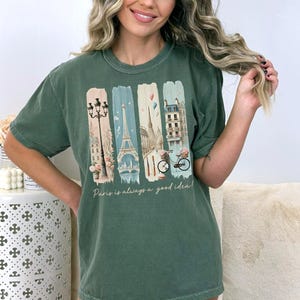 May include: Green t-shirt with a graphic print of Parisian landmarks, including the Eiffel Tower, a street lamp, and a bicycle. The text "Paris is always a good idea" is printed below the graphic.