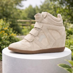 May include: Beige high-top wedge sneakers with three hook-and-loop straps. The sneakers have a suede upper, a brown rubber sole, and a hidden wedge heel. The shoes are displayed on a white pedestal against a blurred green background.