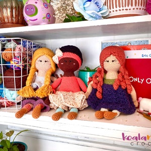May include: Three crocheted dolls sitting on a white shelf. The dolls are wearing different colored dresses and have different hair colors. The doll on the left has blonde hair and a yellow dress, the doll in the middle has brown skin and a pink dress, and the doll on the right has red hair and a purple dress. The dolls are all smiling.