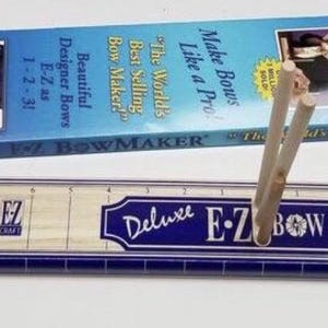 May include: Deluxe E-Z Bow Maker kit with a blue and white design. The kit includes a bow-making tool with wooden dowels and a box with images of bows and text that reads "Make Bows Like a Pro!" and "The World's Best Selling Bow Maker!"