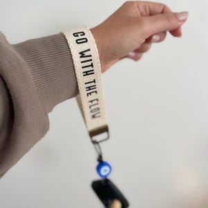 May include: A white canvas wristlet with the text "GO WITH THE FLOW" in black lettering. A blue evil eye charm is attached to the wristlet.