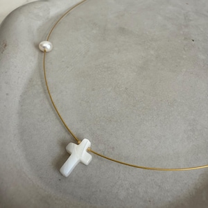 May include: A delicate gold-tone necklace featuring a white cross pendant and a single pearl. The cross pendant has a marbled appearance. The necklace is displayed on a light gray surface.